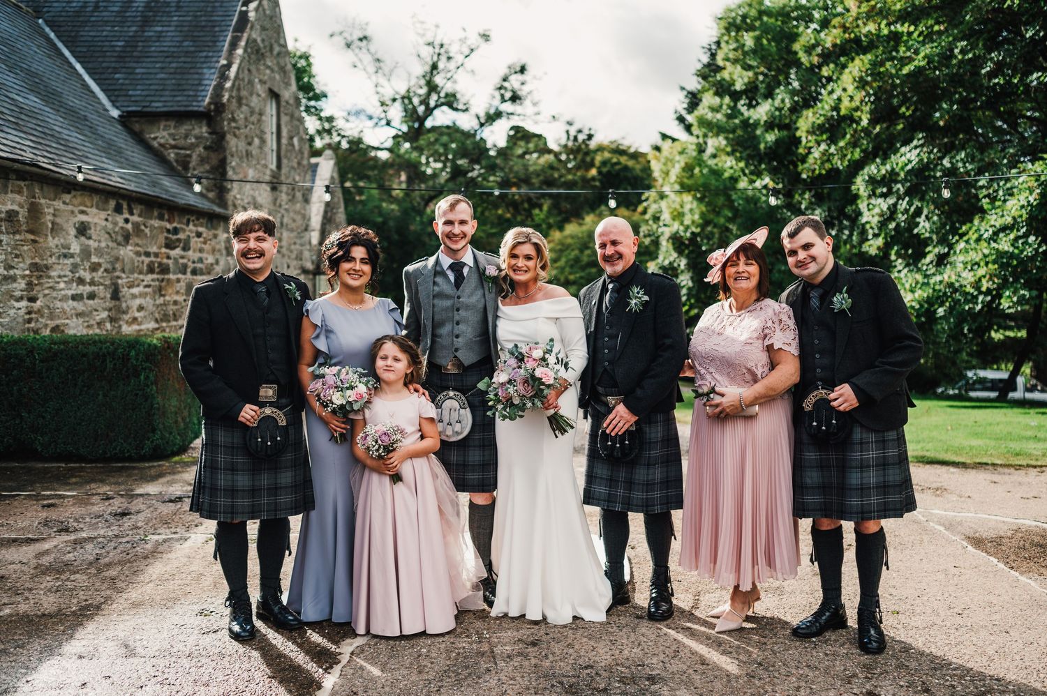 Wedding party in black suits and pink dresses poses together outdoors at a stone building.