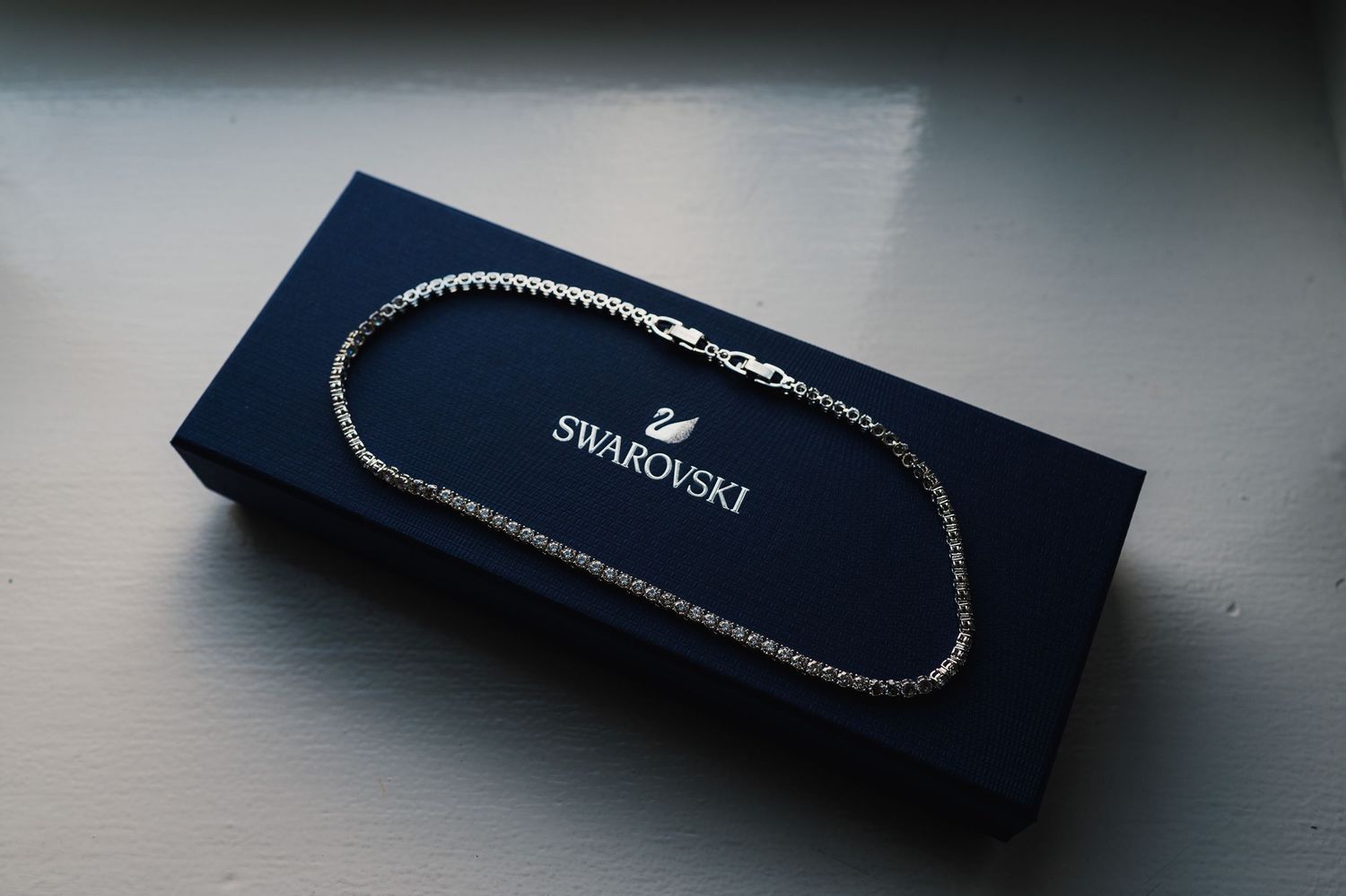A silver Swarovski crystal necklace displayed with its dark blue retail box in a product photography setup.