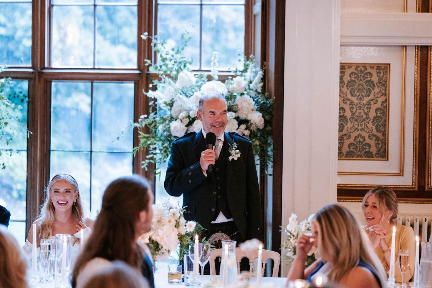 Elegant wedding reception venue with large windows, floral arrangements and seated guests.