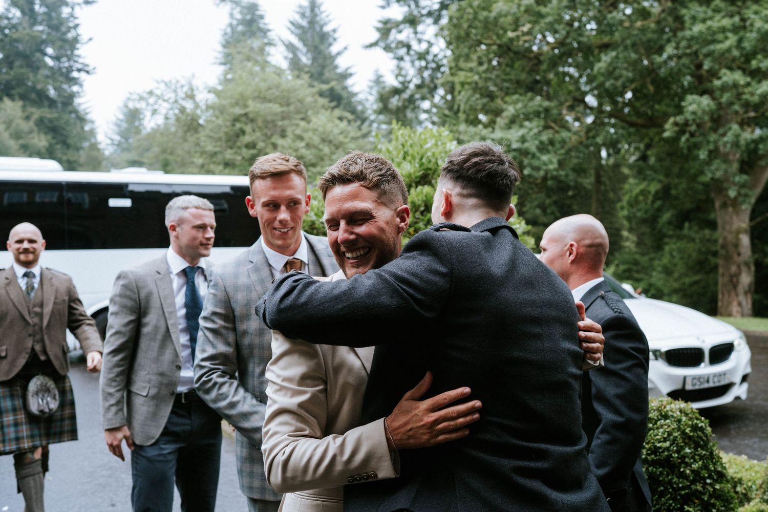 Groomsmen share laughs and hugs outside in a candid moment at a wedding celebration.