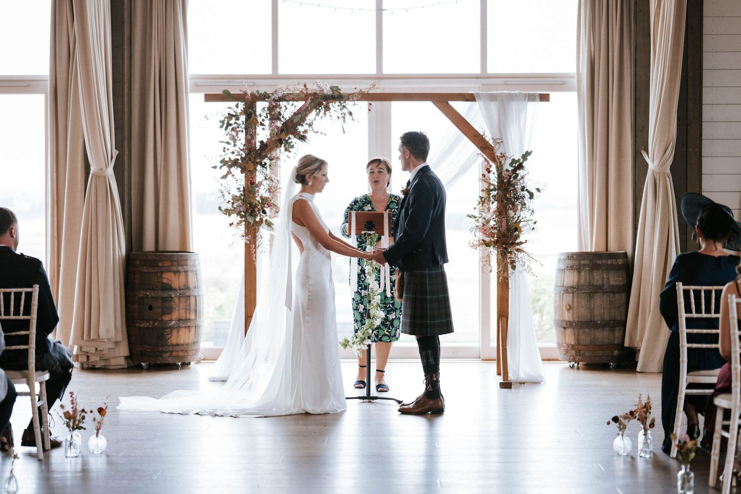Wedding ceremony setup in bright indoor venue with large windows and white curtains.