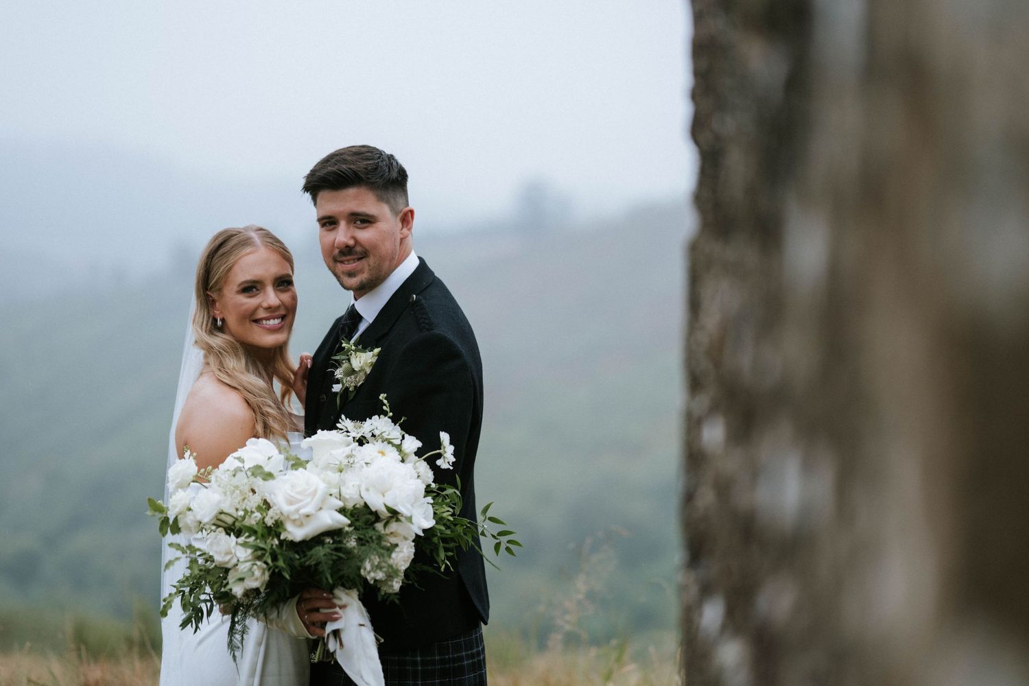 Newlywed couple embraces during an outdoor photo session with misty mountains in the background.