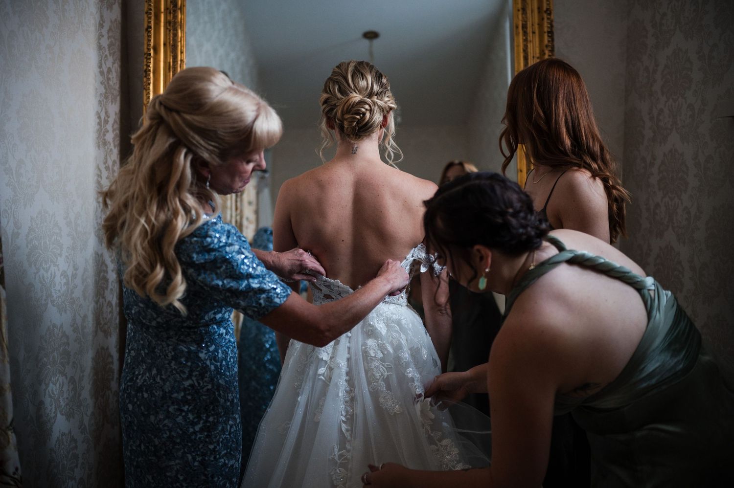 Bridesmaids help with final dress preparations in a dimly lit hallway before the wedding ceremony.