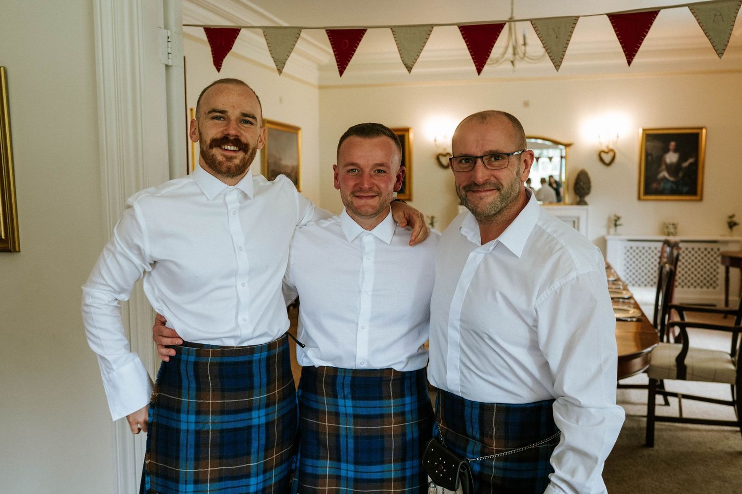 Wedding party members in white shirts and blue tartan kilts posing together.