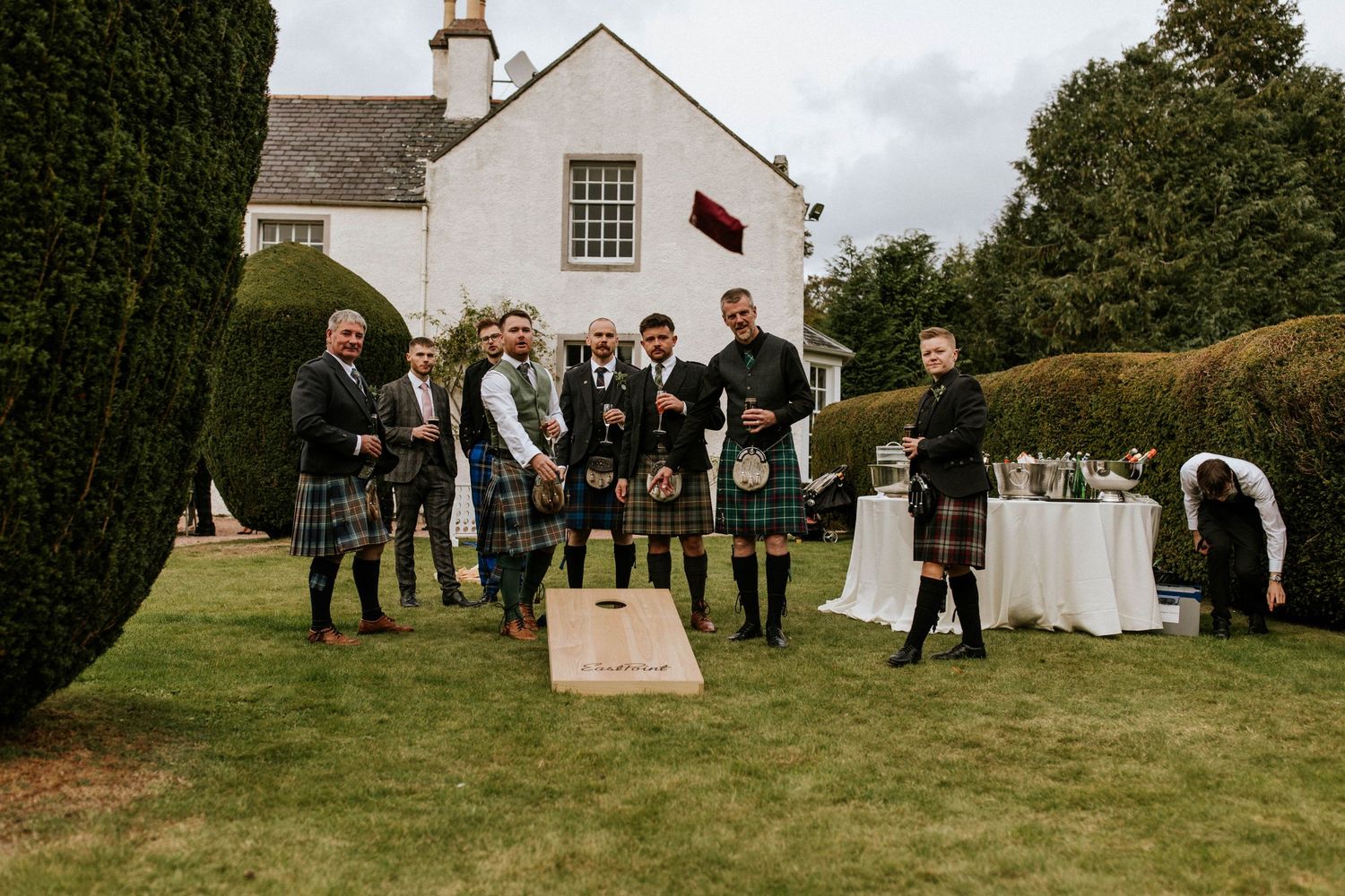 A group in traditional Scottish kilts gathers on a lawn during a wedding celebration.