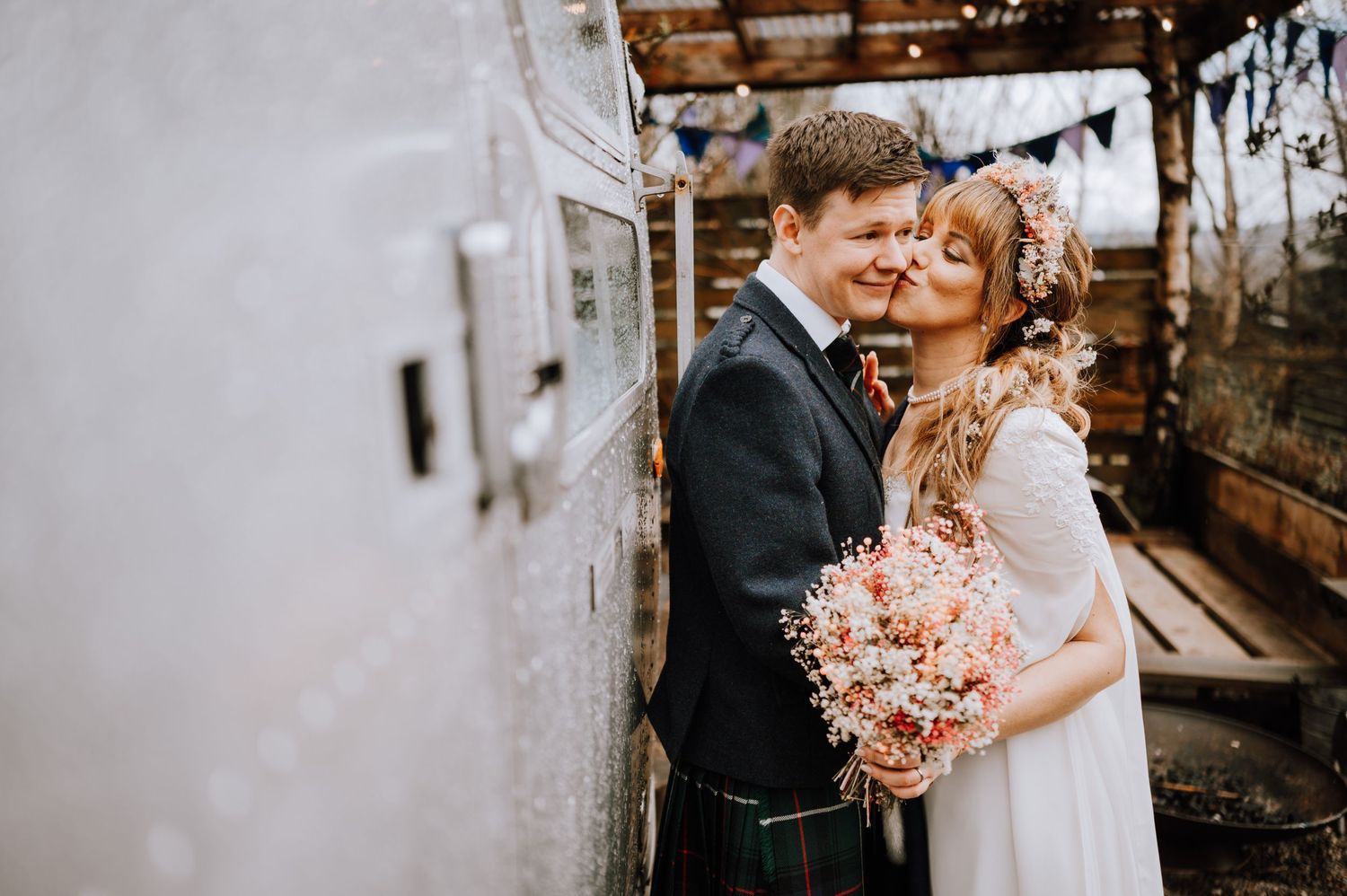 Tender moment between newlyweds against rustic wall with bride holding pink floral bouquet.