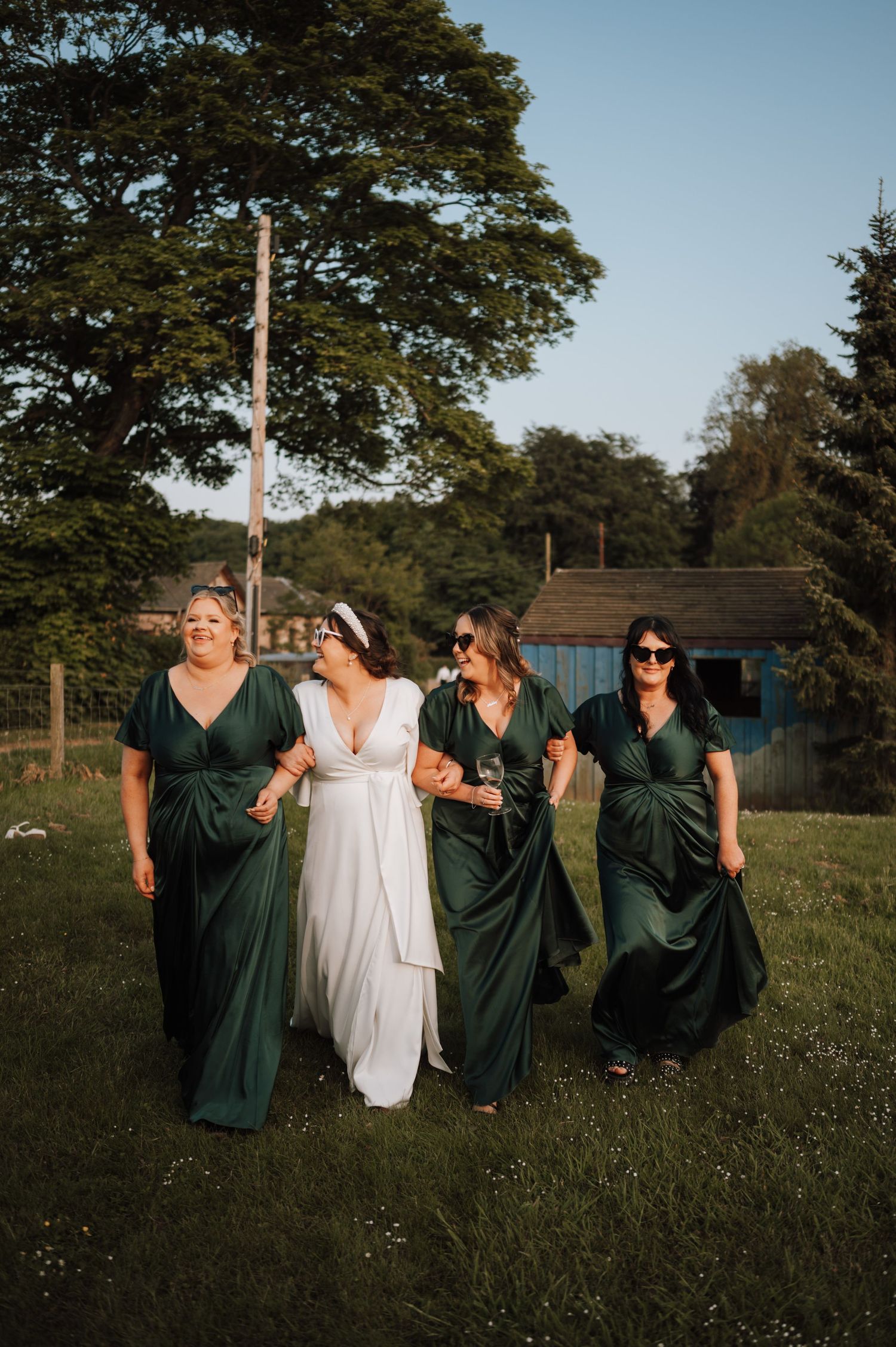 Bridal party in elegant green dresses walks through garden setting at sunset.