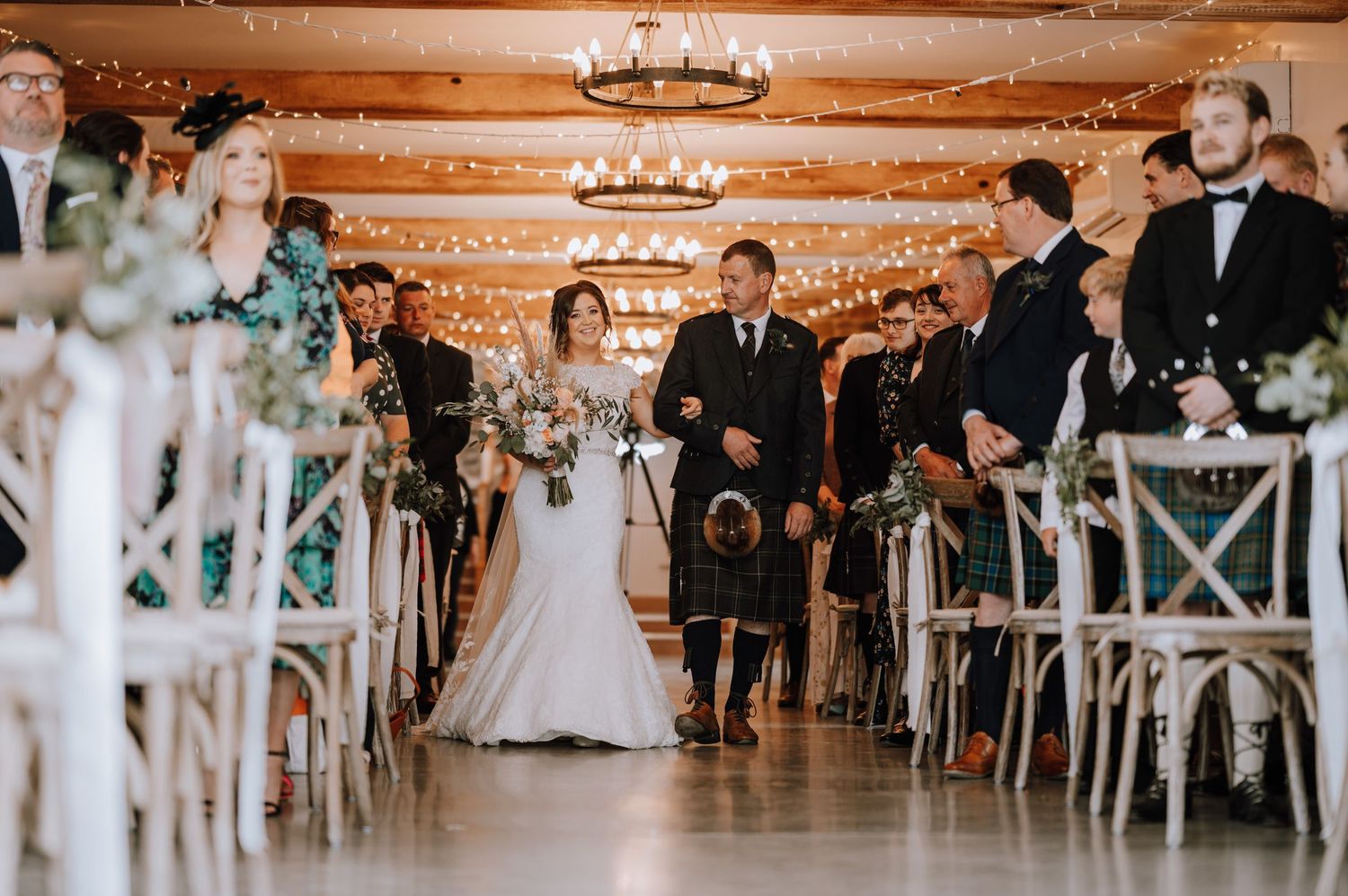 Ceremony processional in a barn with wooden beams and string lights creating a warm, romantic atmosphere.