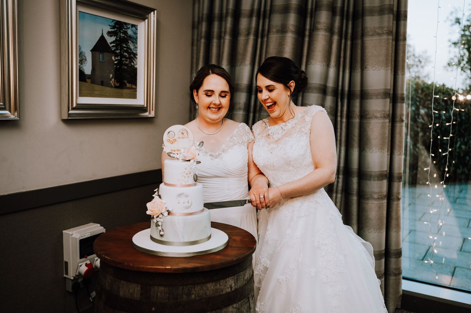 Two brides admire their white tiered wedding cake together in an elegant indoor setting.
