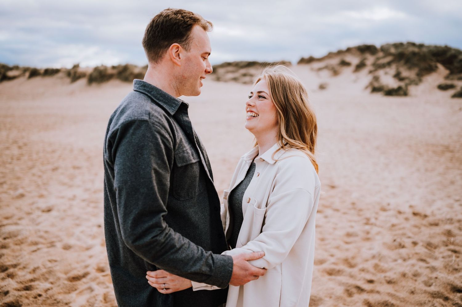 Couple sharing tender moment while standing on beach with cloudy backdrop.