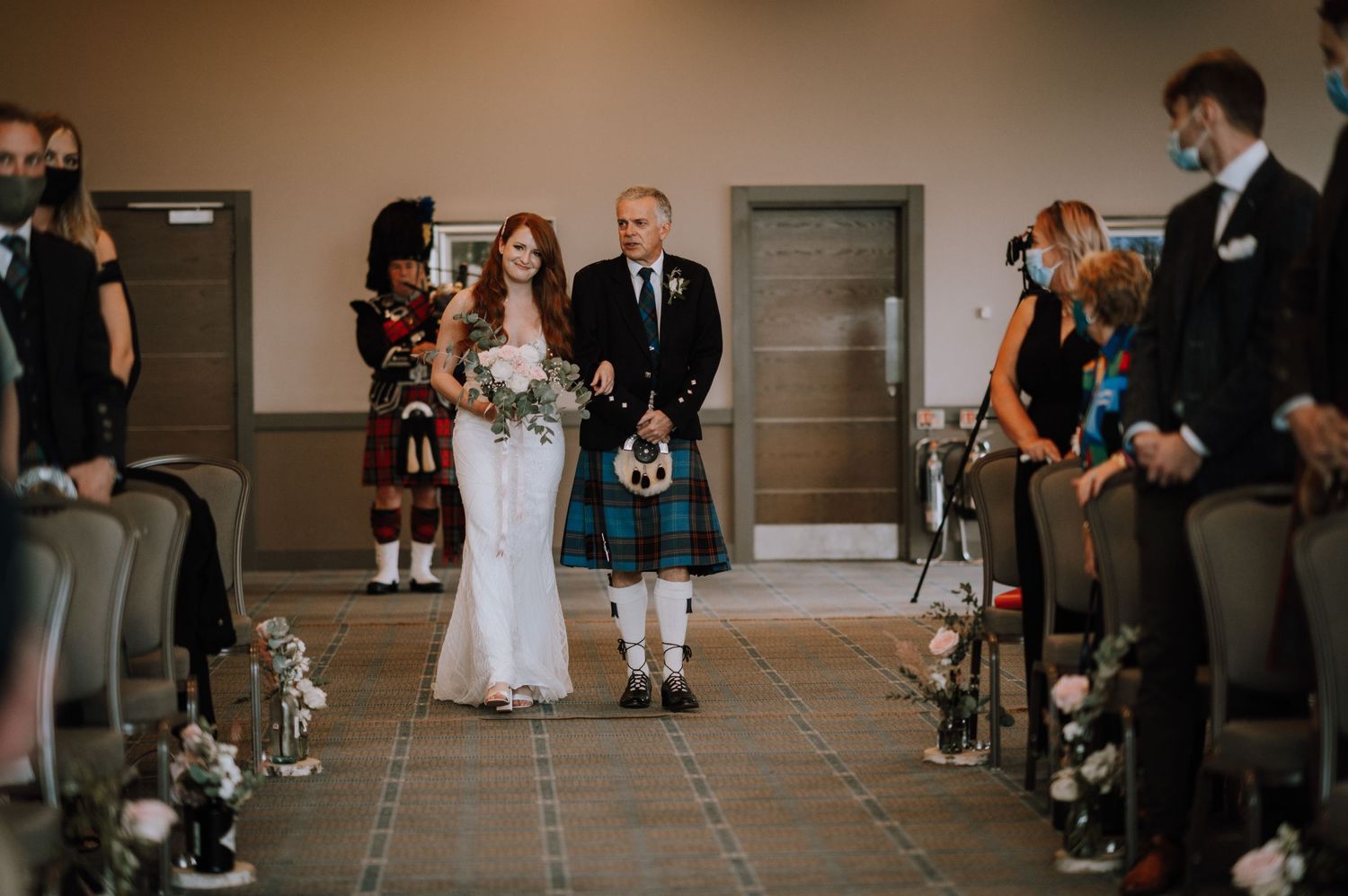 Wedding processional in venue with Scottish kilts and white dress walking down aisle.