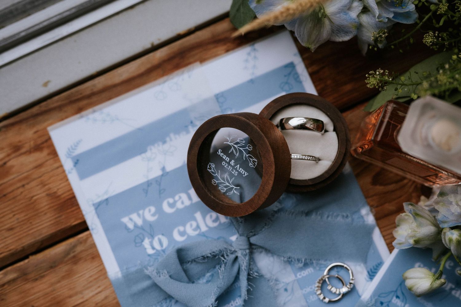 Wedding rings and invitation details arranged on a wooden surface with floral accents.