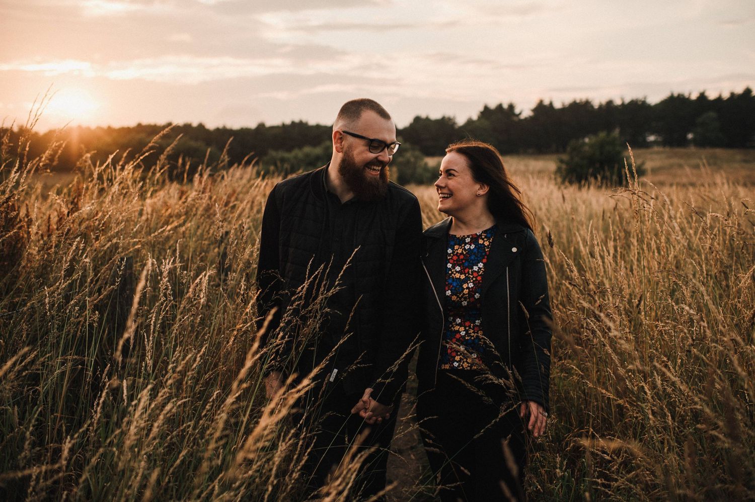 A joyful couple shares a laugh while standing together in a sunlit meadow at dusk.