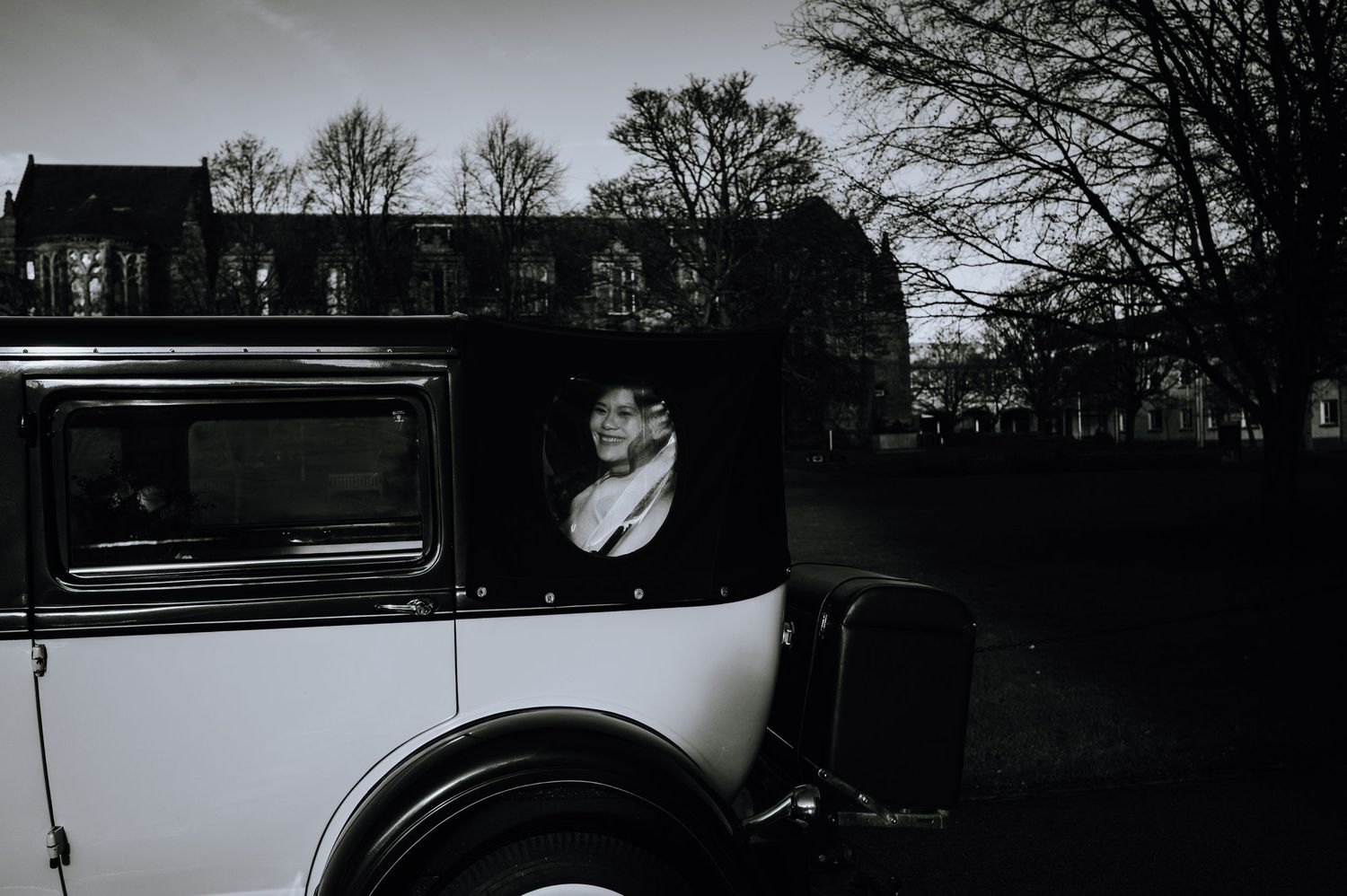 A vintage black and white photo shows a classic car with a passenger looking out the window.
