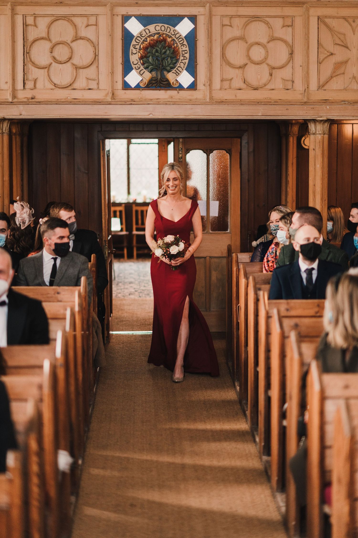 A bridesmaid in burgundy dress walks down church aisle while masked guests look on.