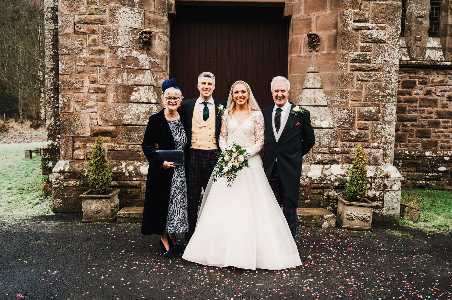 Small wedding group poses in front of rustic stone church entrance with traditional wooden door.