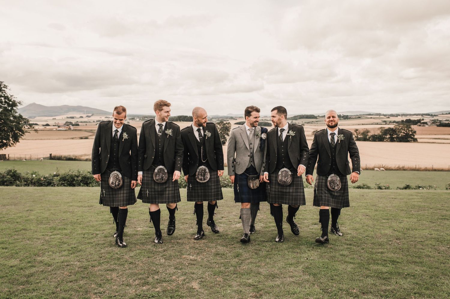 Wedding party in traditional Scottish kilts walking across green hillside landscape.