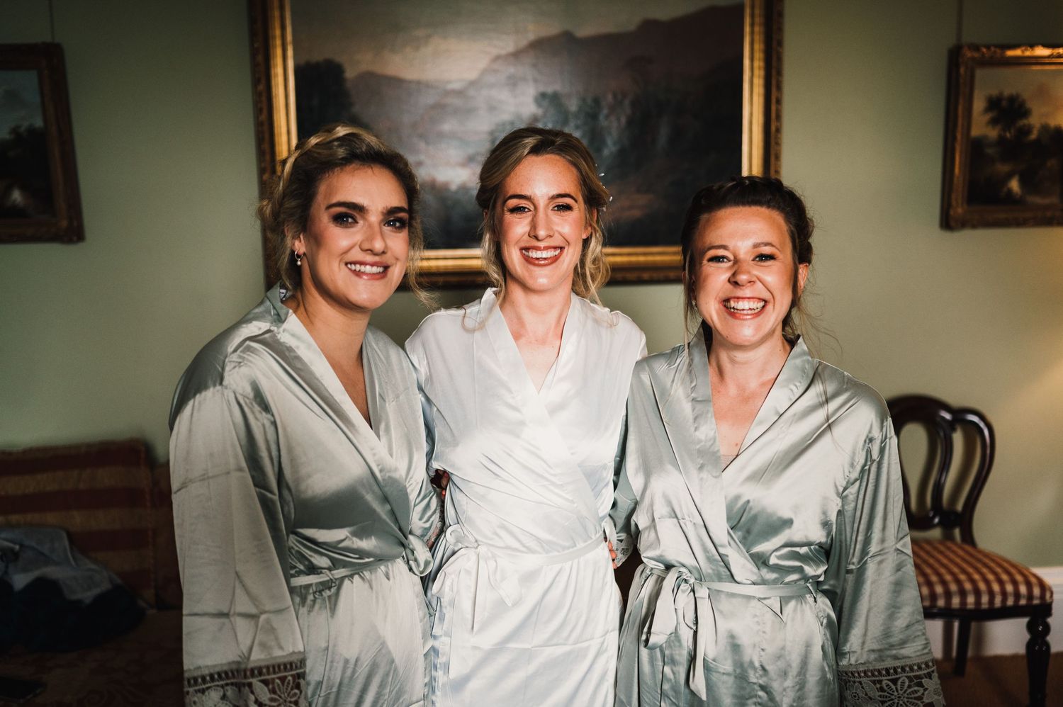 Three people pose together wearing matching satin robes during wedding preparations.