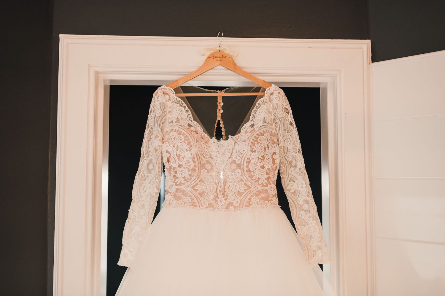 A white wedding dress with lace bodice and long sleeves hangs on a wooden hanger against a white door.
