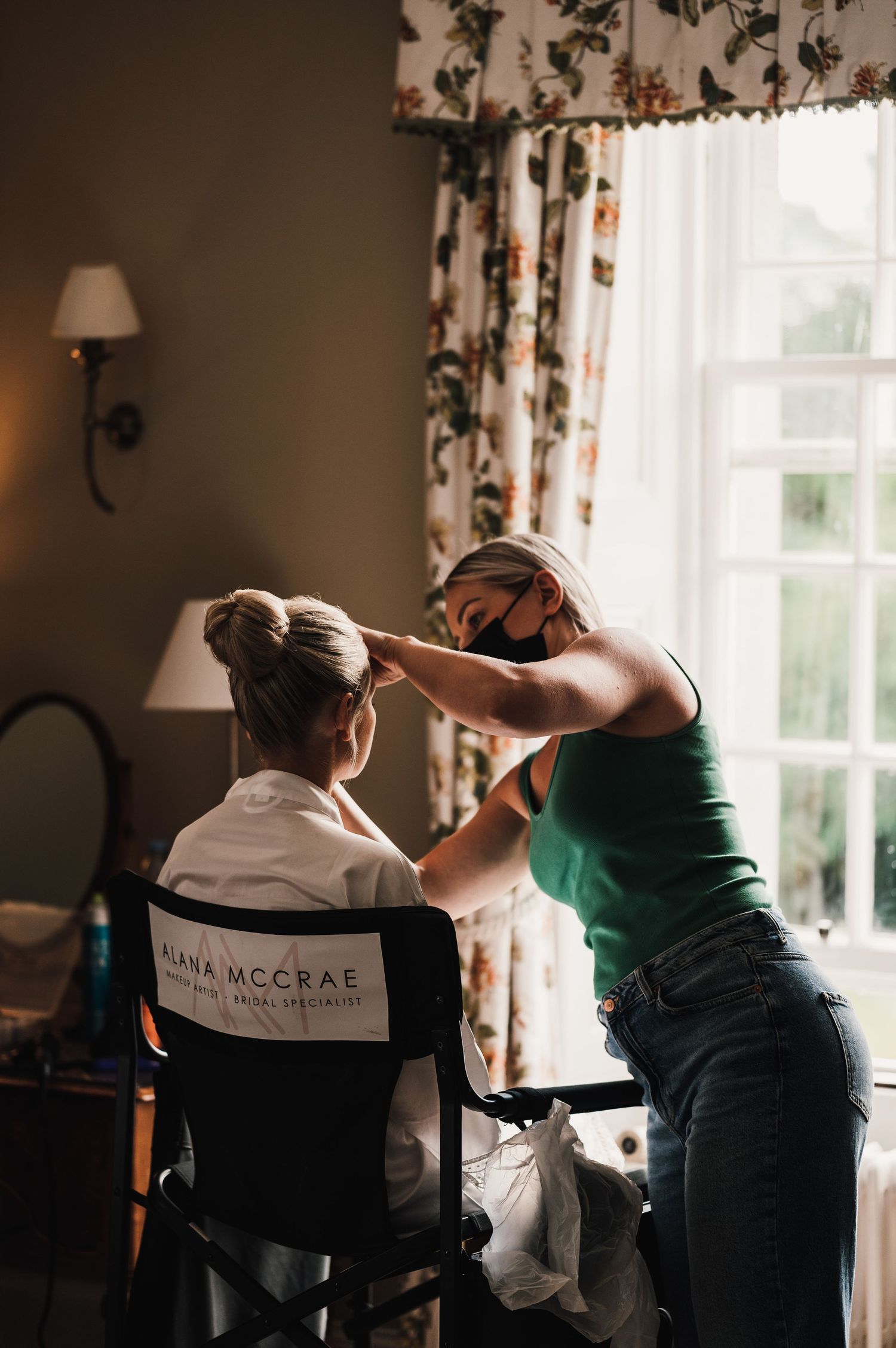 A stylist works on hair preparation in a chair near a window with floral curtains.
