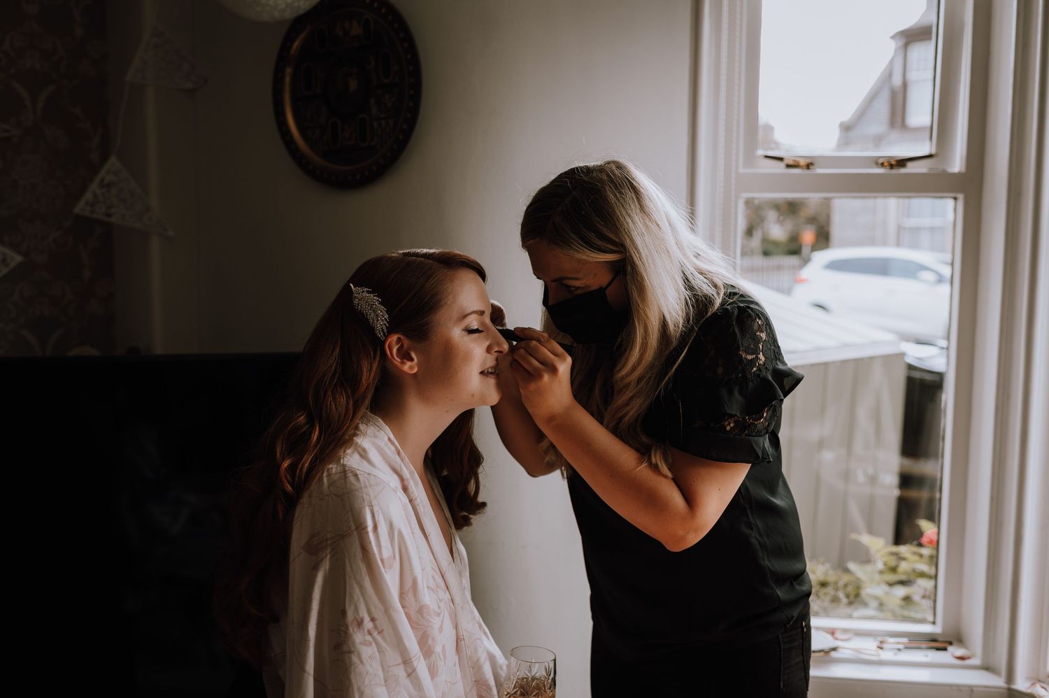 A makeup application session taking place by a bright window in a home setting.