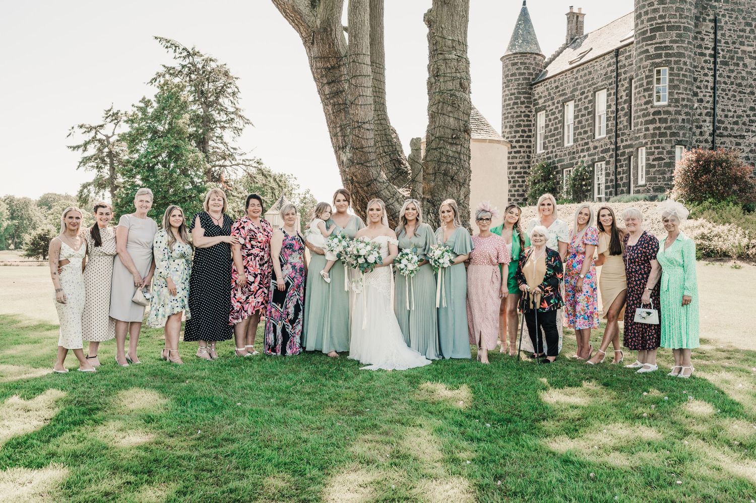 Large wedding group photo featuring guests in pastel dresses and formal wear on castle grounds.