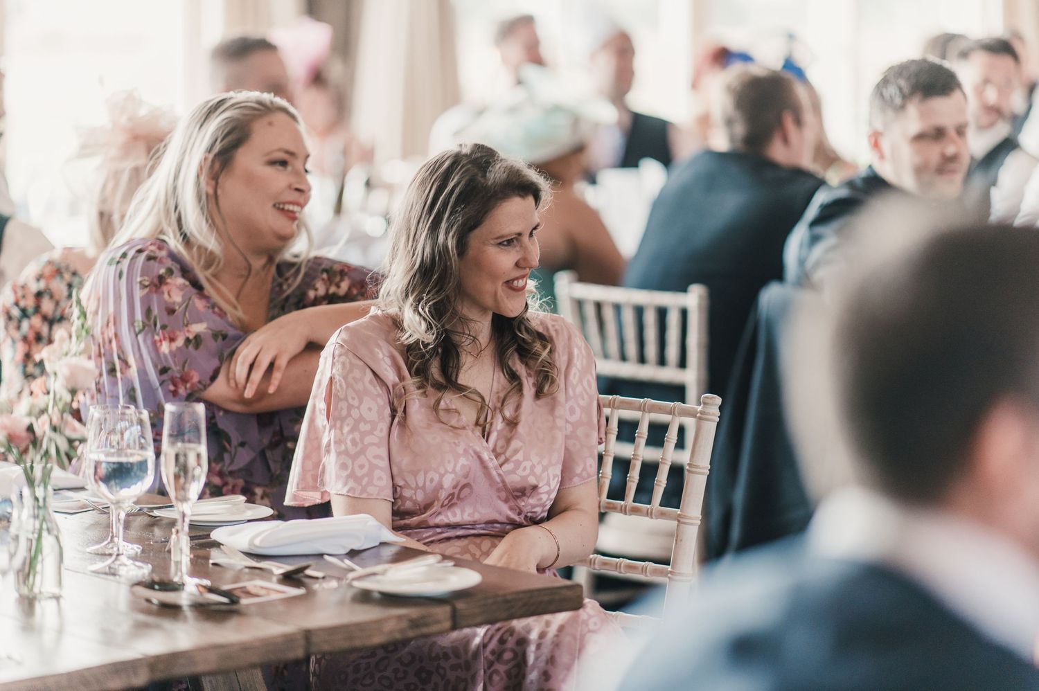 Wedding guests in formal attire laugh and smile during a reception at an elegant venue.
