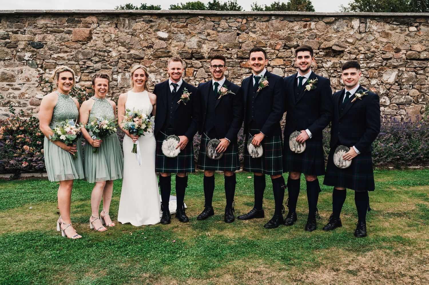 Wedding party in formal attire with kilts and bridesmaid dresses pose together against rustic stone wall.