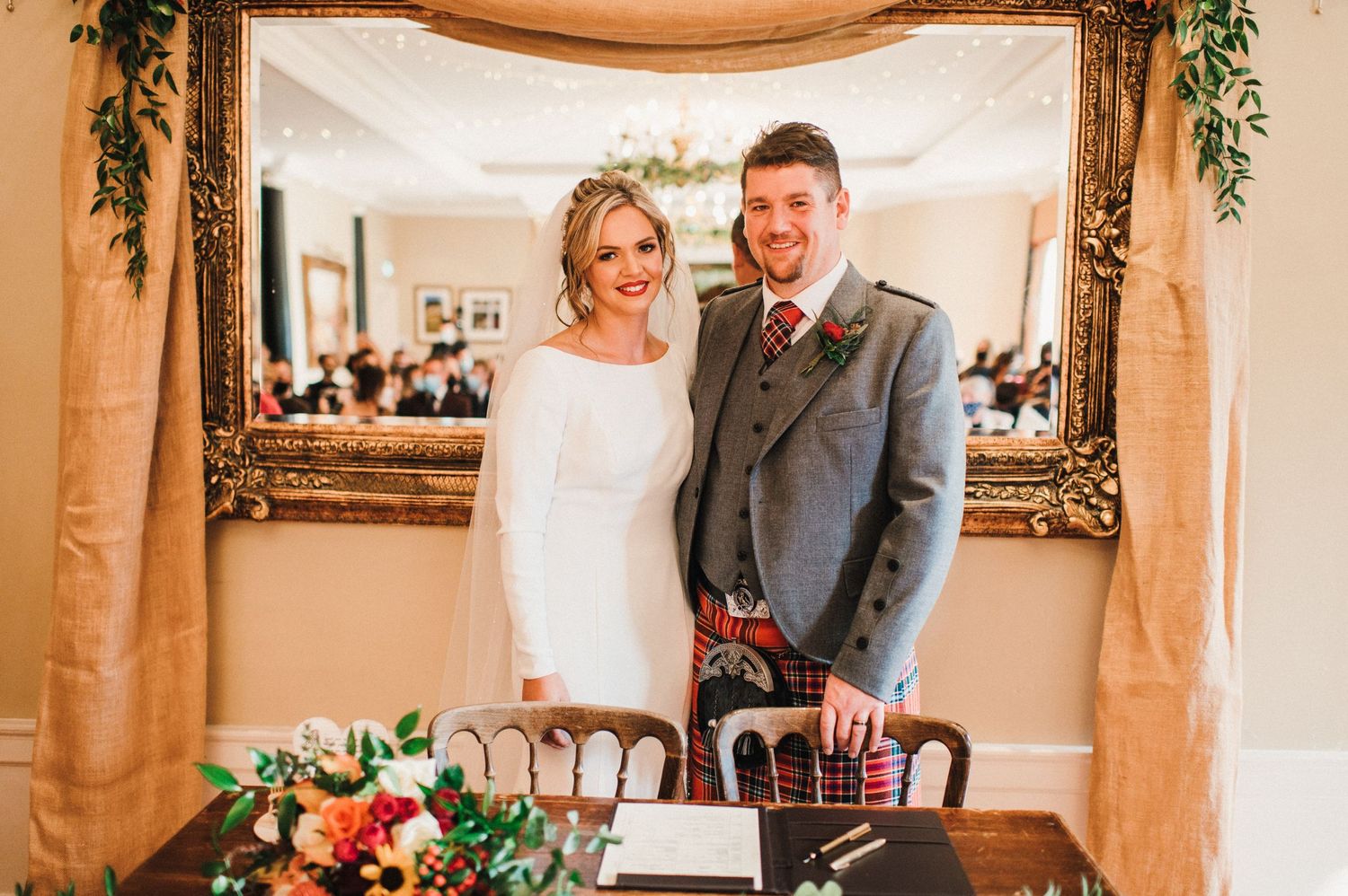 Wedding couple stands under elegant draped arch with lush floral arrangements.