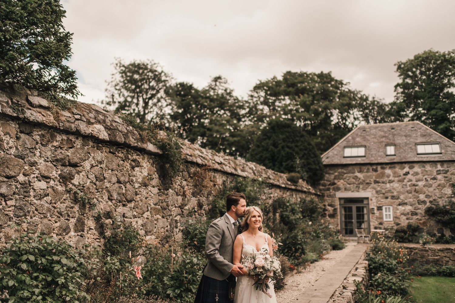 A romantic wedding couple stands by an old stone wall and garden path leading to a rustic stone building.