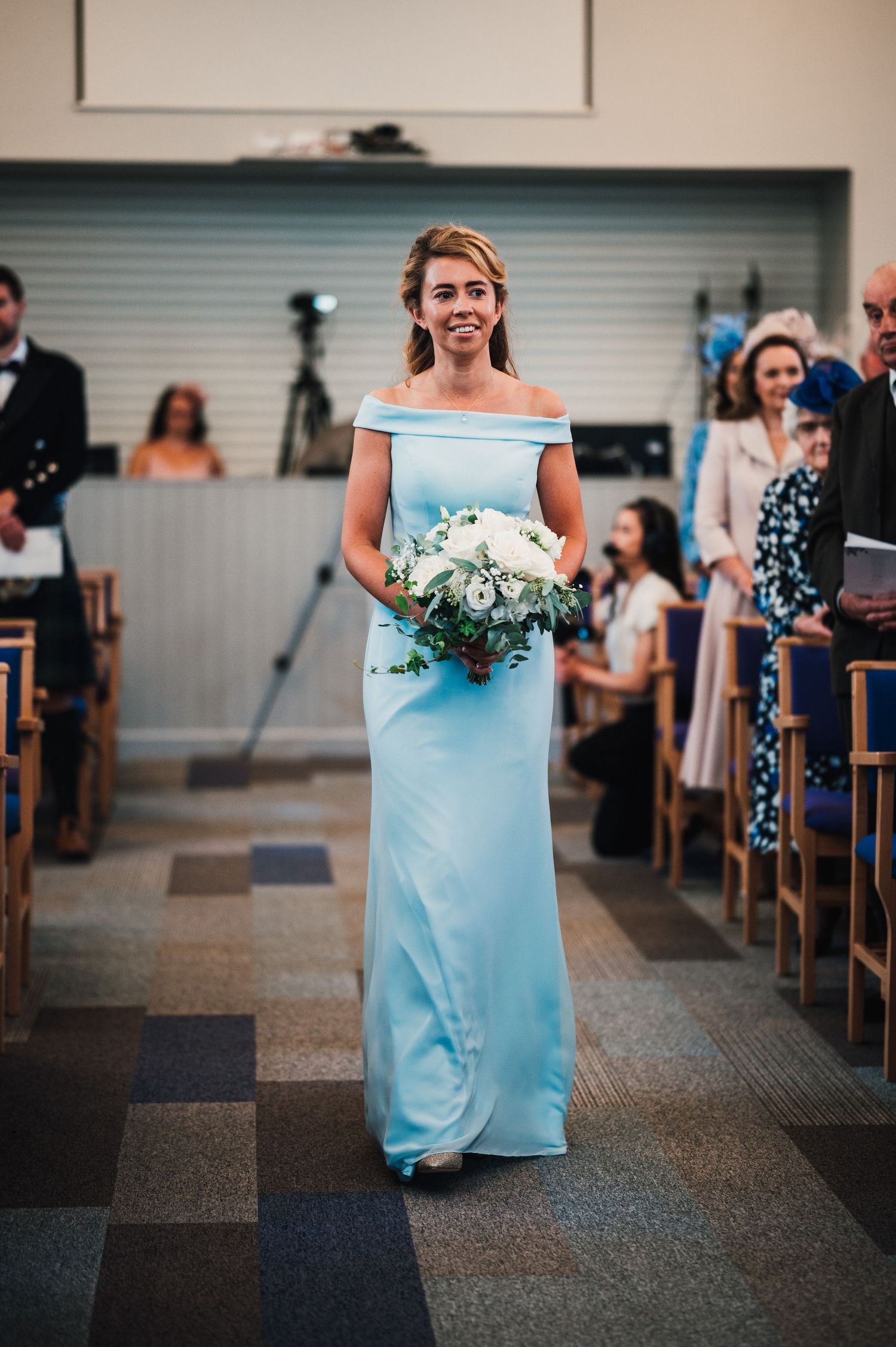 Bridesmaid in light blue off-shoulder gown walks down church aisle with white flower bouquet.