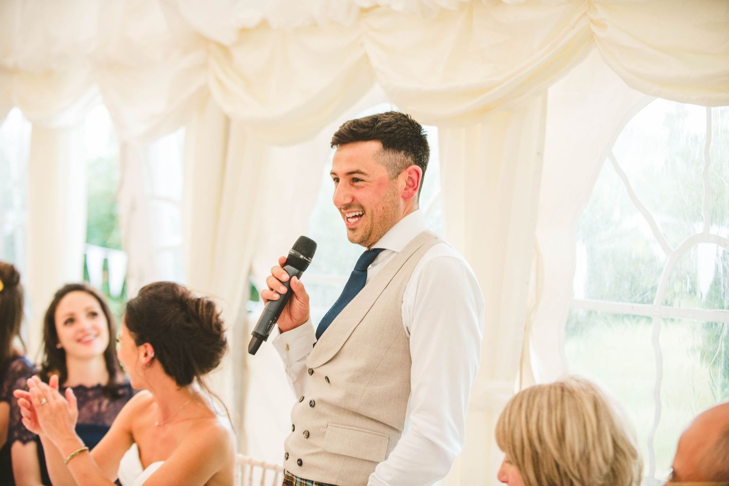 Wedding reception speaker with microphone entertaining guests in white marquee tent.