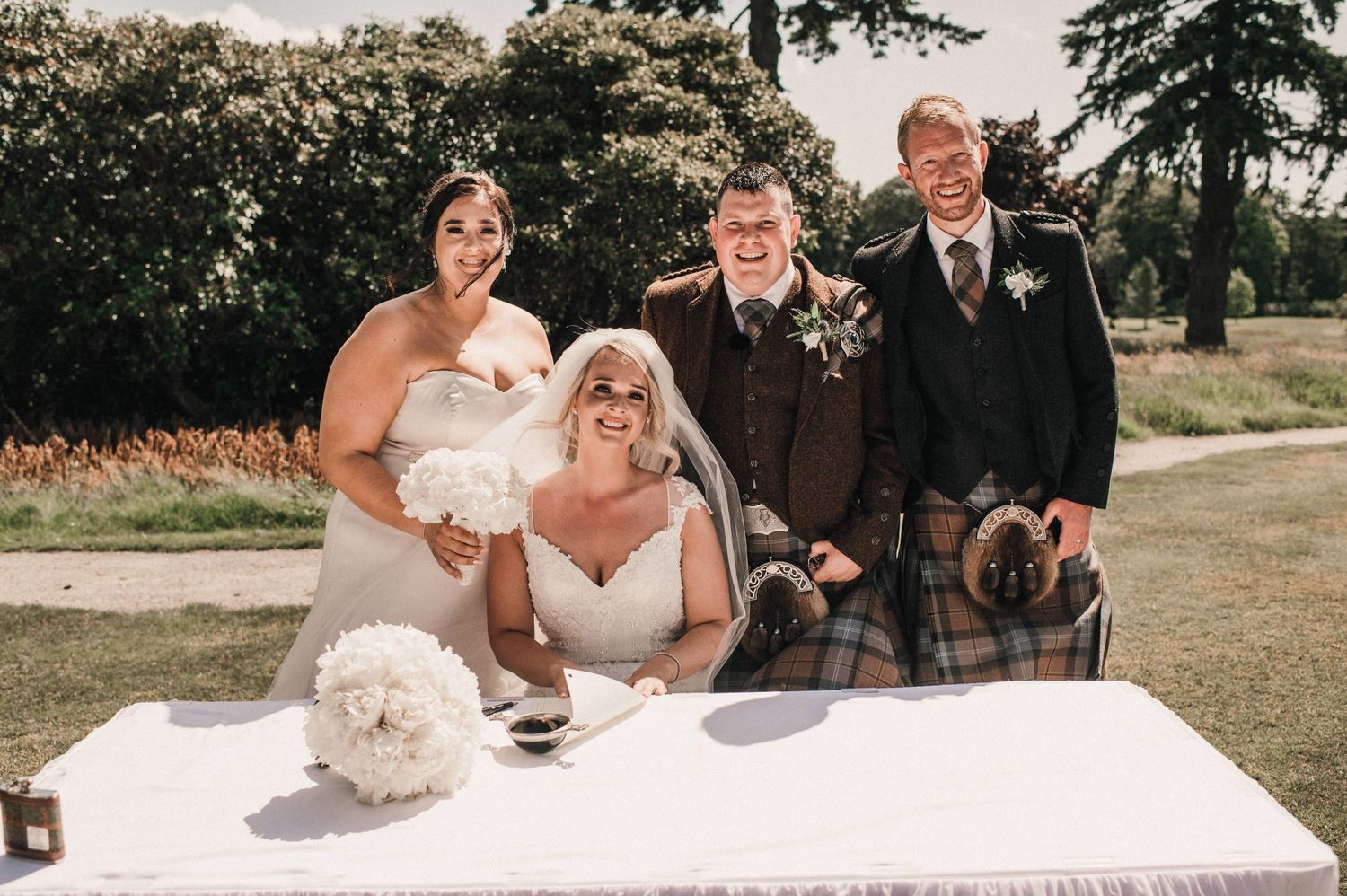 Wedding party gathered at signing table with traditional Scottish kilts and white floral decorations.