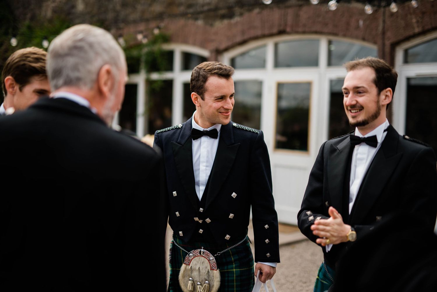 Men in formal black tie and traditional Scottish kilts conversing at an event.