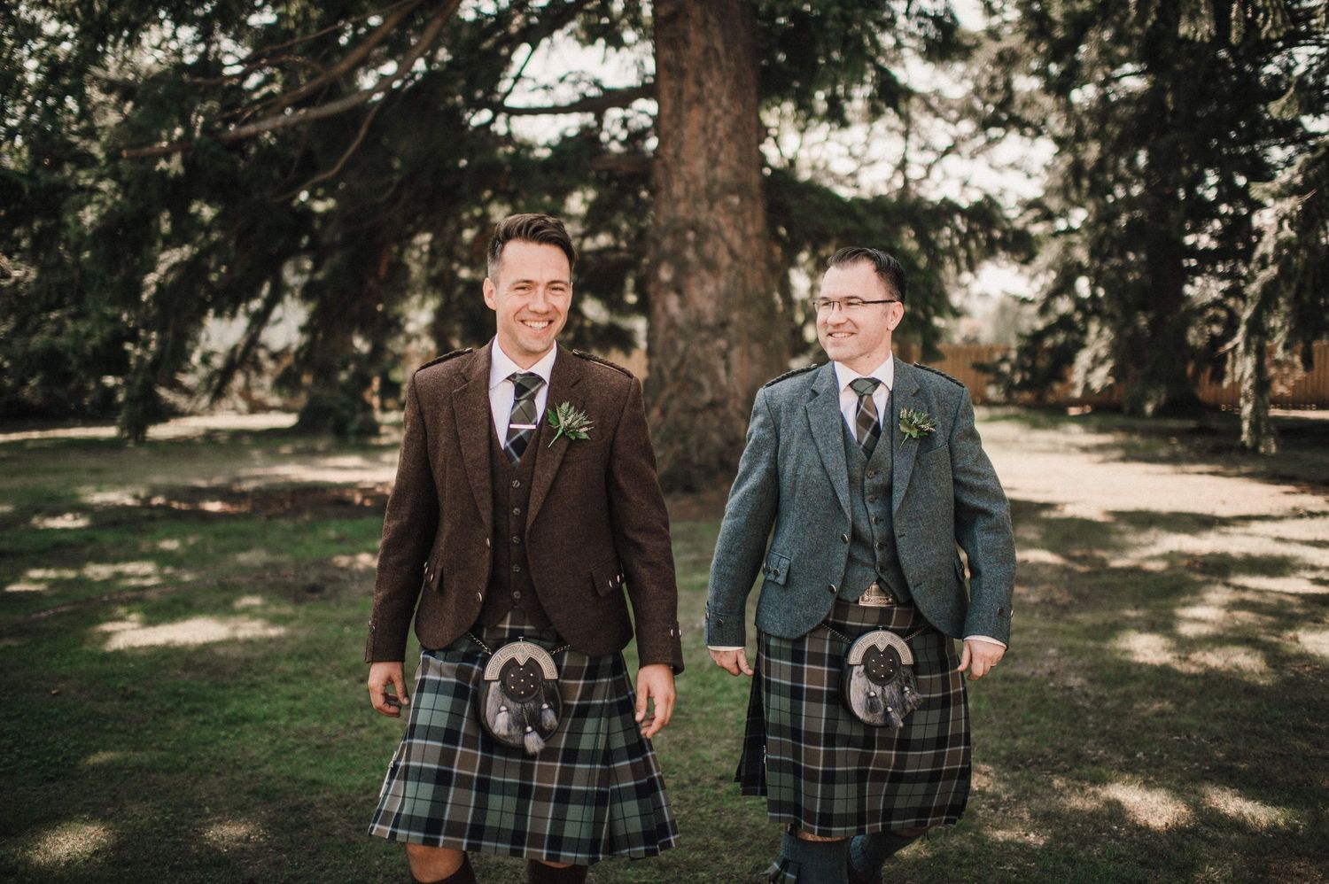 Two people wearing matching kilts and tweed jackets pose together in a garden setting.