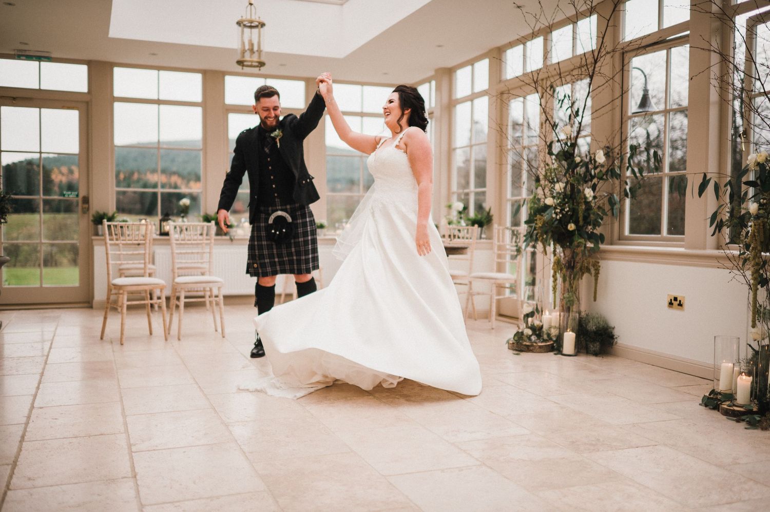Wedding couple dances joyfully in bright conservatory with large windows.
