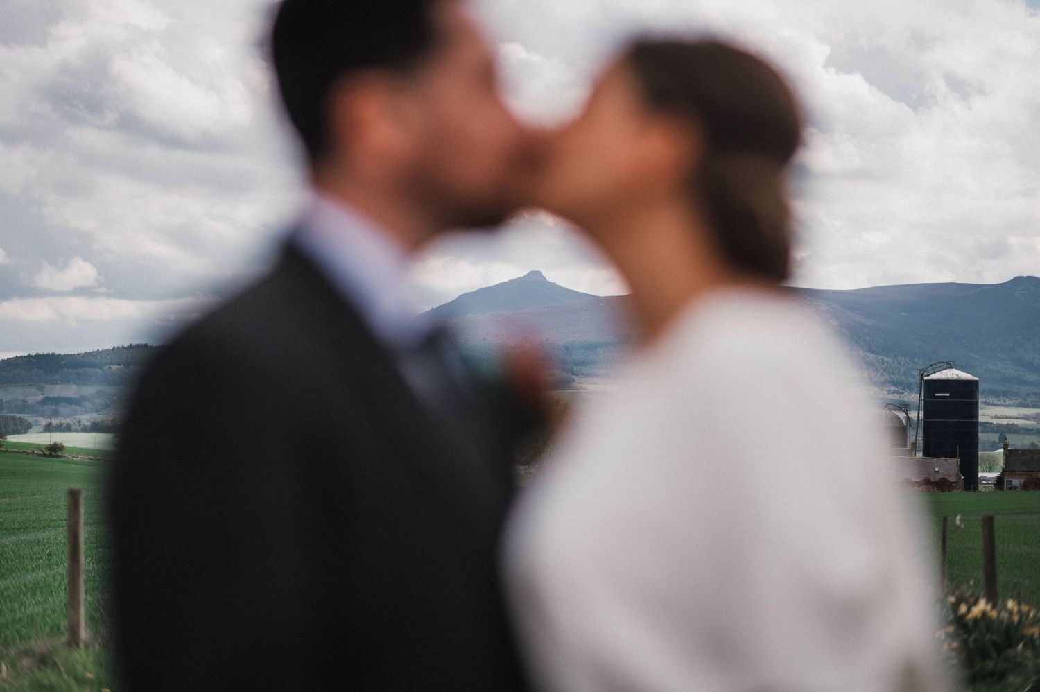 A romantic wedding couple shares a kiss against a scenic mountain backdrop in soft focus.
