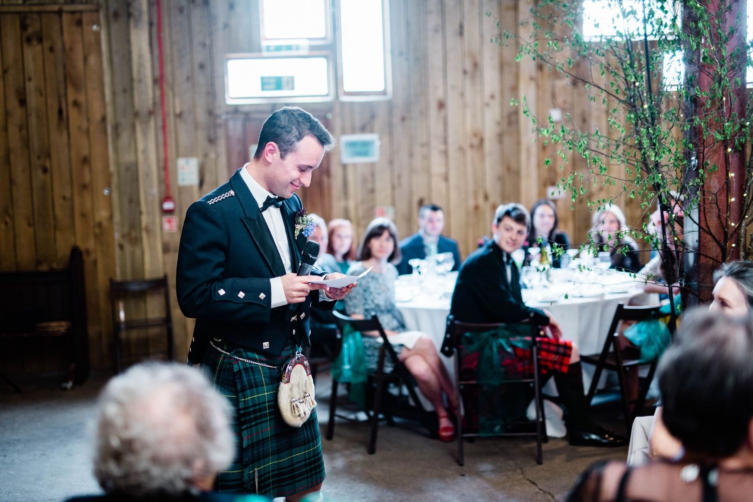 Speaker in traditional Scottish attire addresses guests at wedding reception with wooden decor.