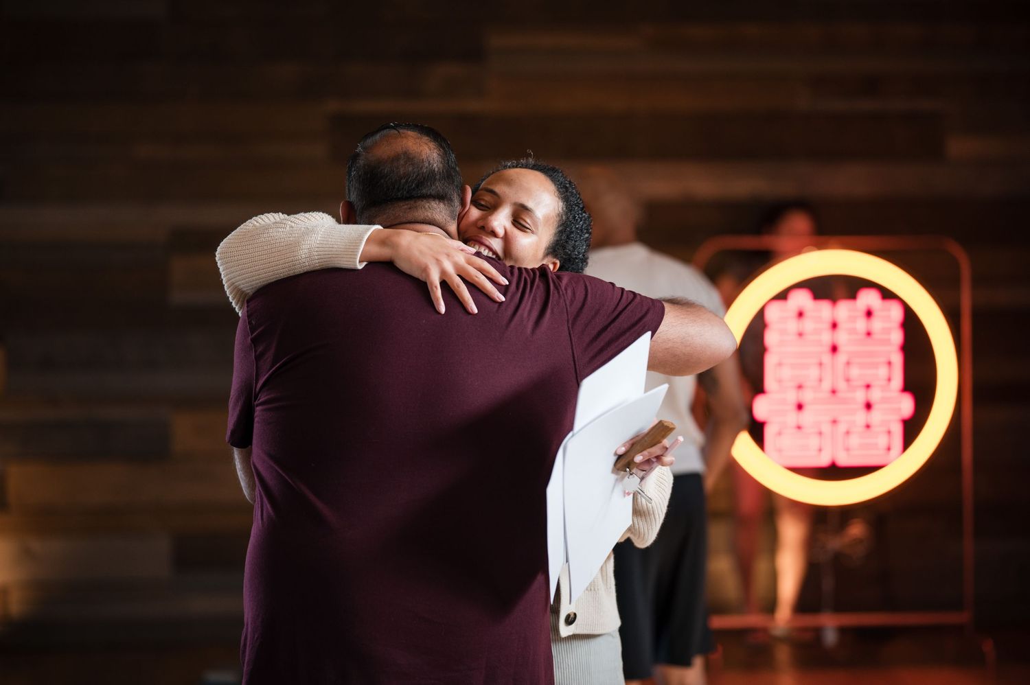 Two people share an emotional embrace on a dance floor with a glowing neon sign in the background.