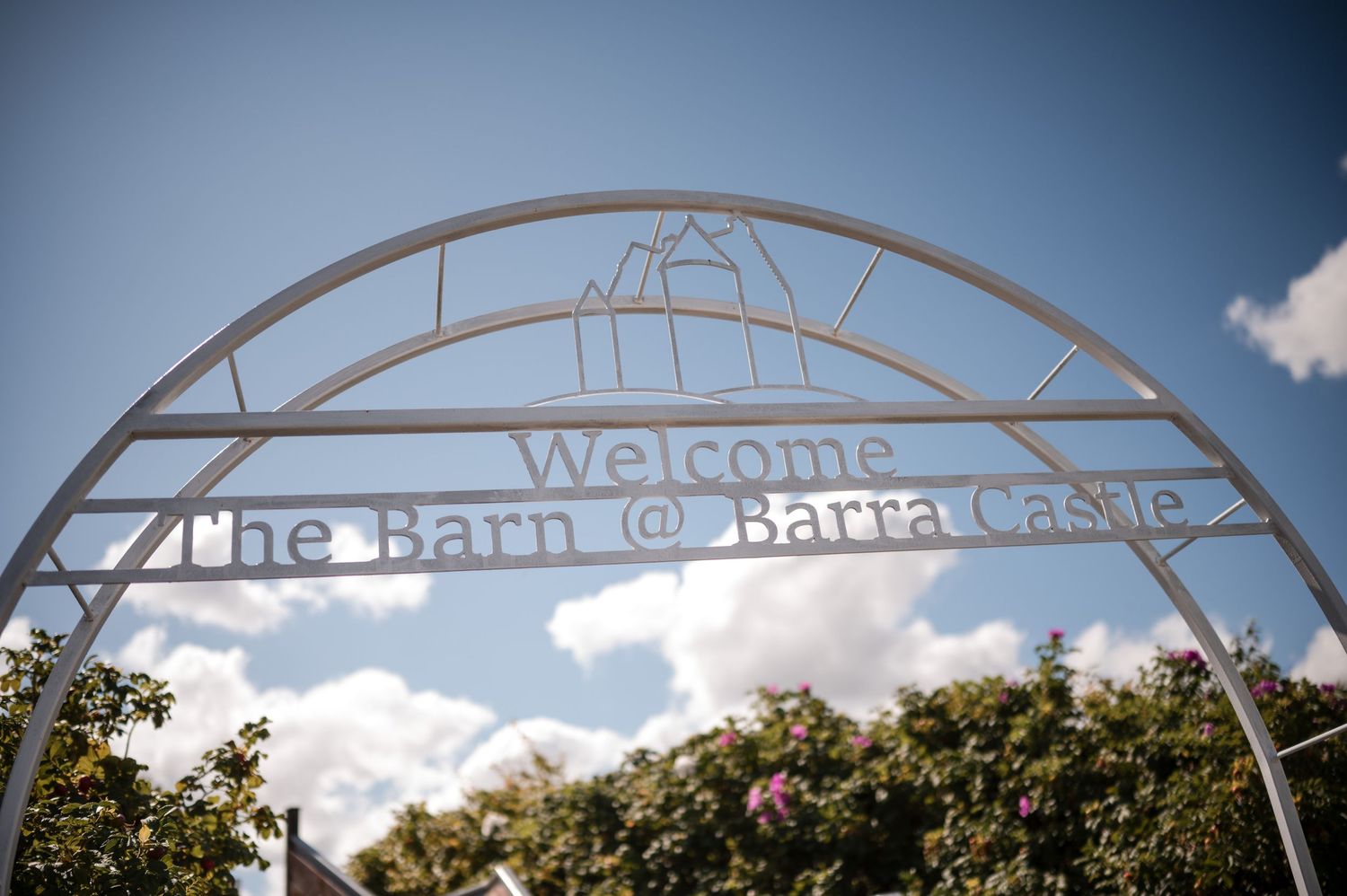 White metal archway entrance with decorative text spans across a garden path under blue skies.