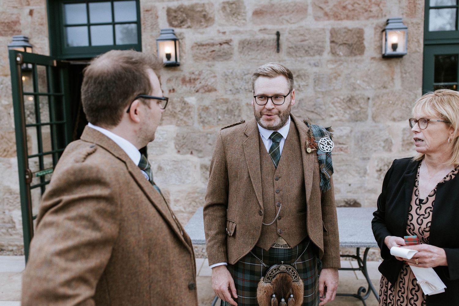 Three guests in formal attire having a conversation outside a stone building with wall-mounted lanterns.