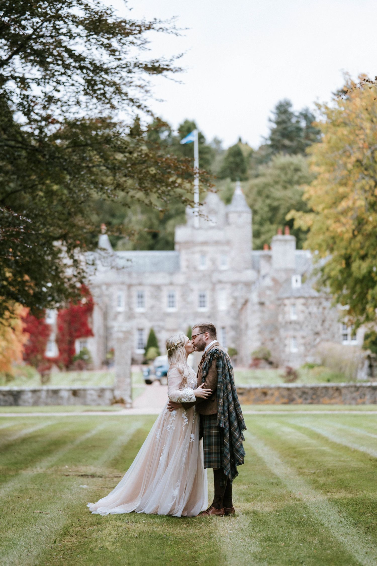 A wedding couple poses in front of a majestic stone castle with autumn trees in the background.