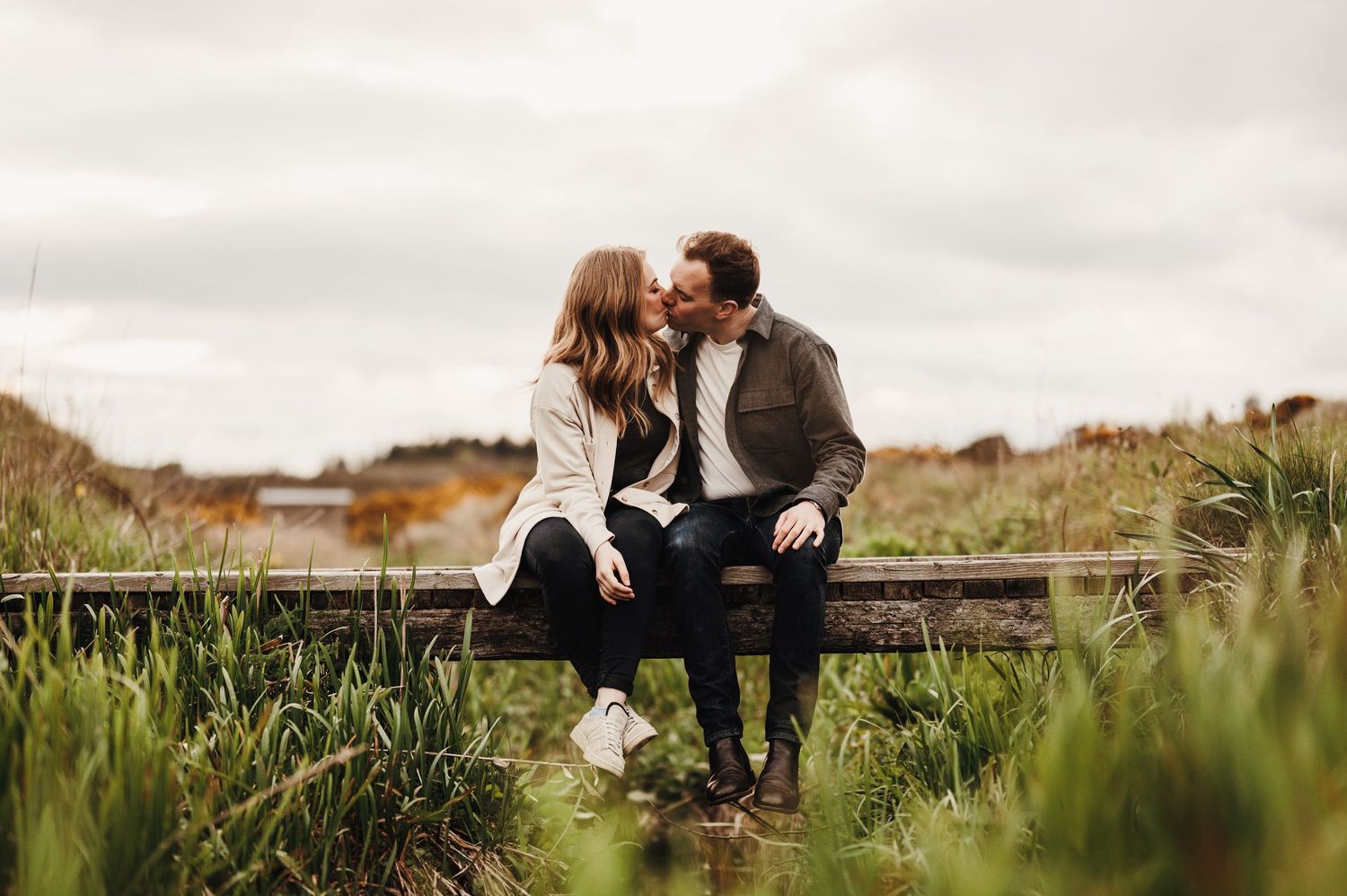A couple sits together on a wooden boardwalk surrounded by tall grass in warm autumn lighting.