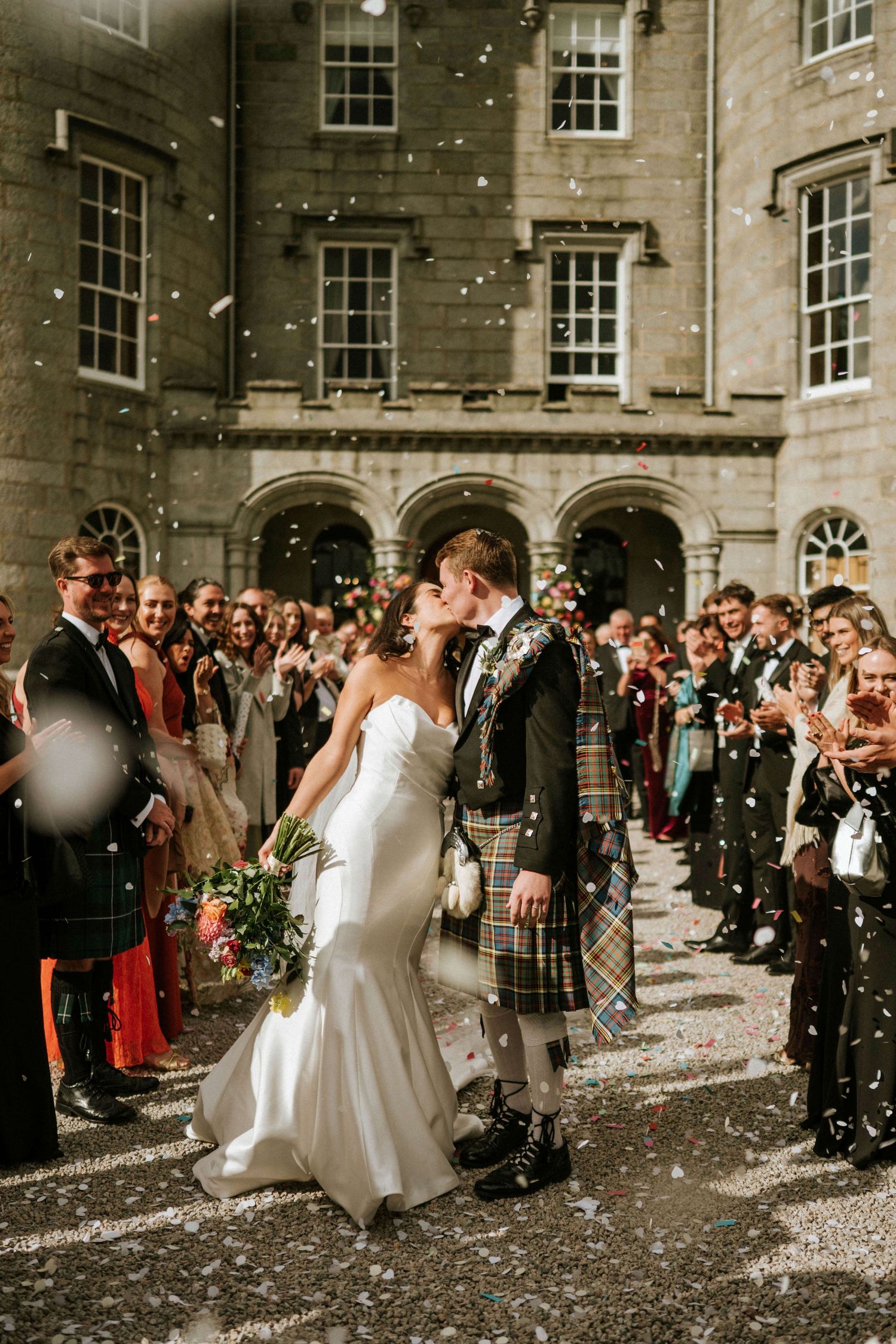 Joyful wedding celebration outside stone building with confetti and gathered guests.