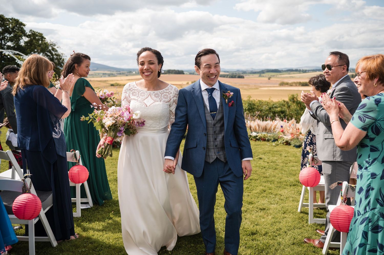 Wedding guests mingle and celebrate outdoors on a grassy hillside with scenic countryside views in the background.