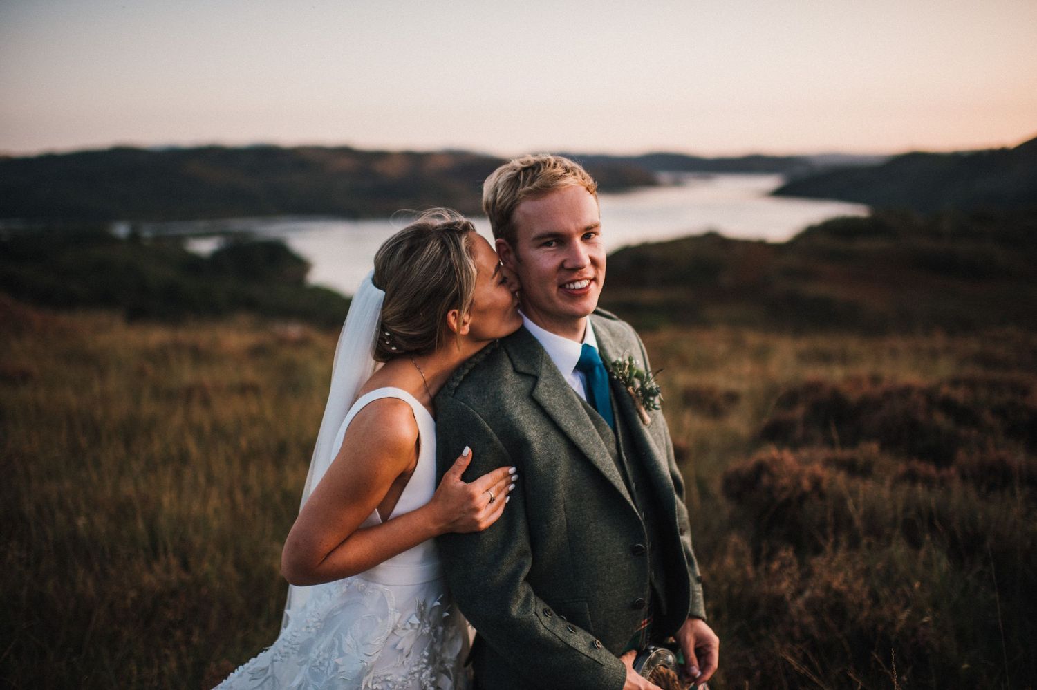 Couple in wedding attire embraces during sunset at scenic overlook with water and mountains in background.