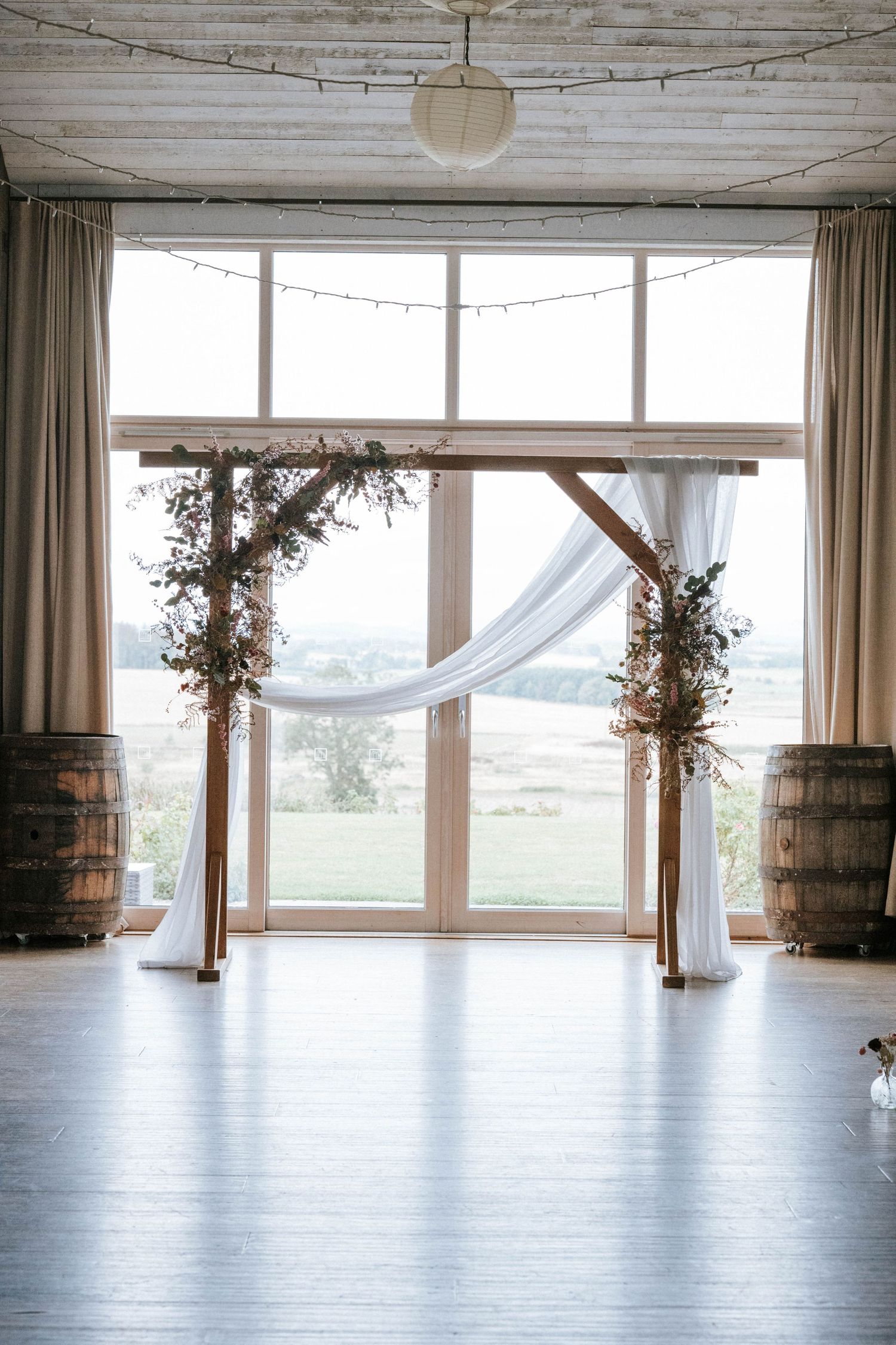 Elegant wedding venue space with rustic wooden barrels and white drapes framing large windows overlooking scenic views.