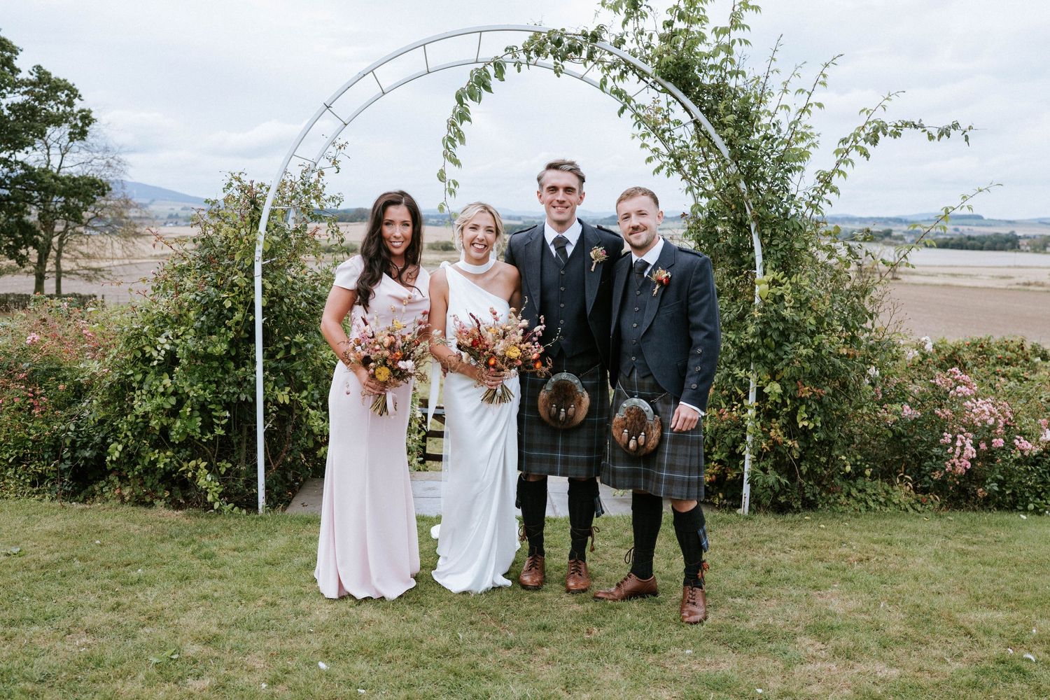 Wedding party members stand together for formal portraits in an outdoor garden setting.