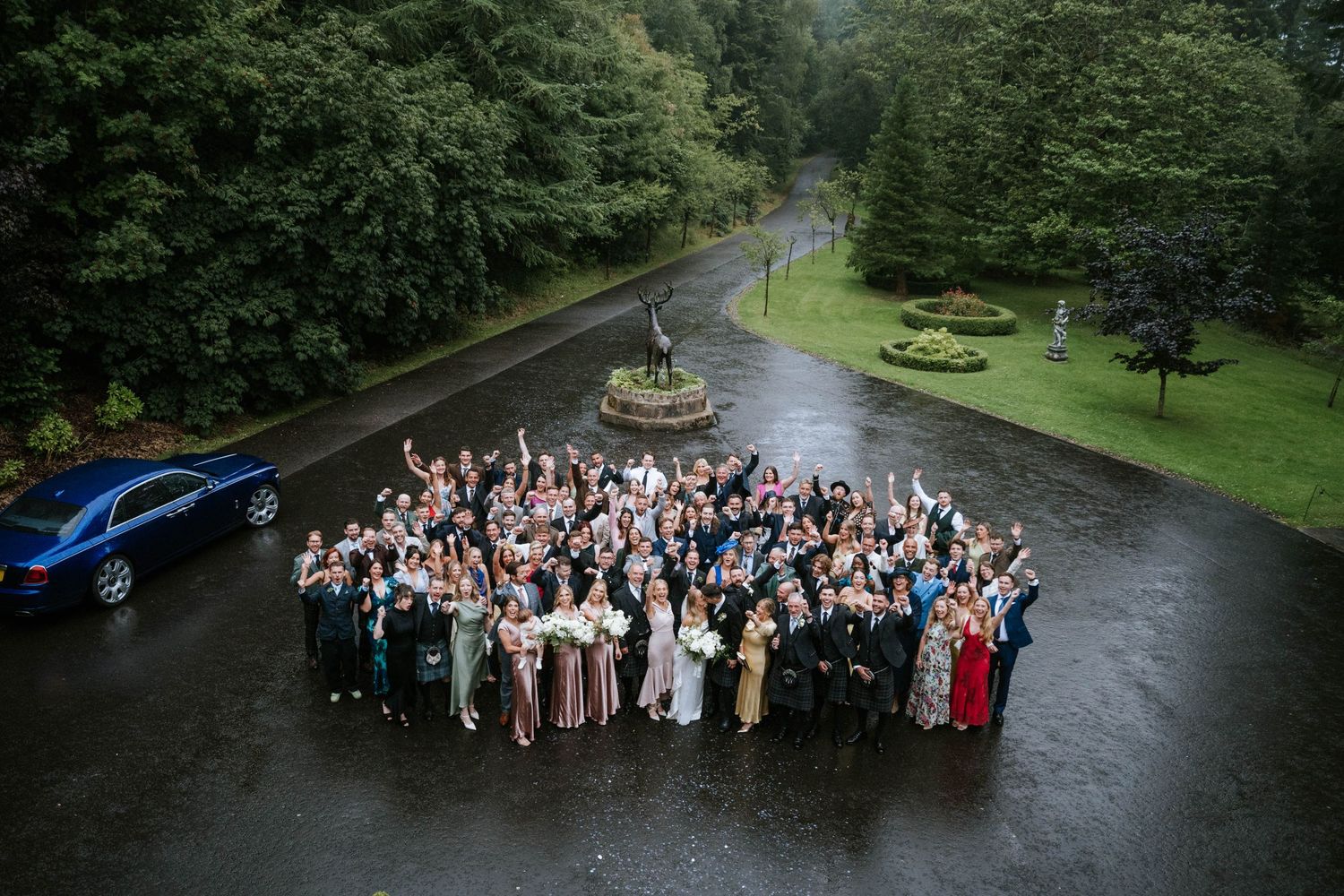Large wedding party group poses outdoors on a misty path surrounded by lush greenery.