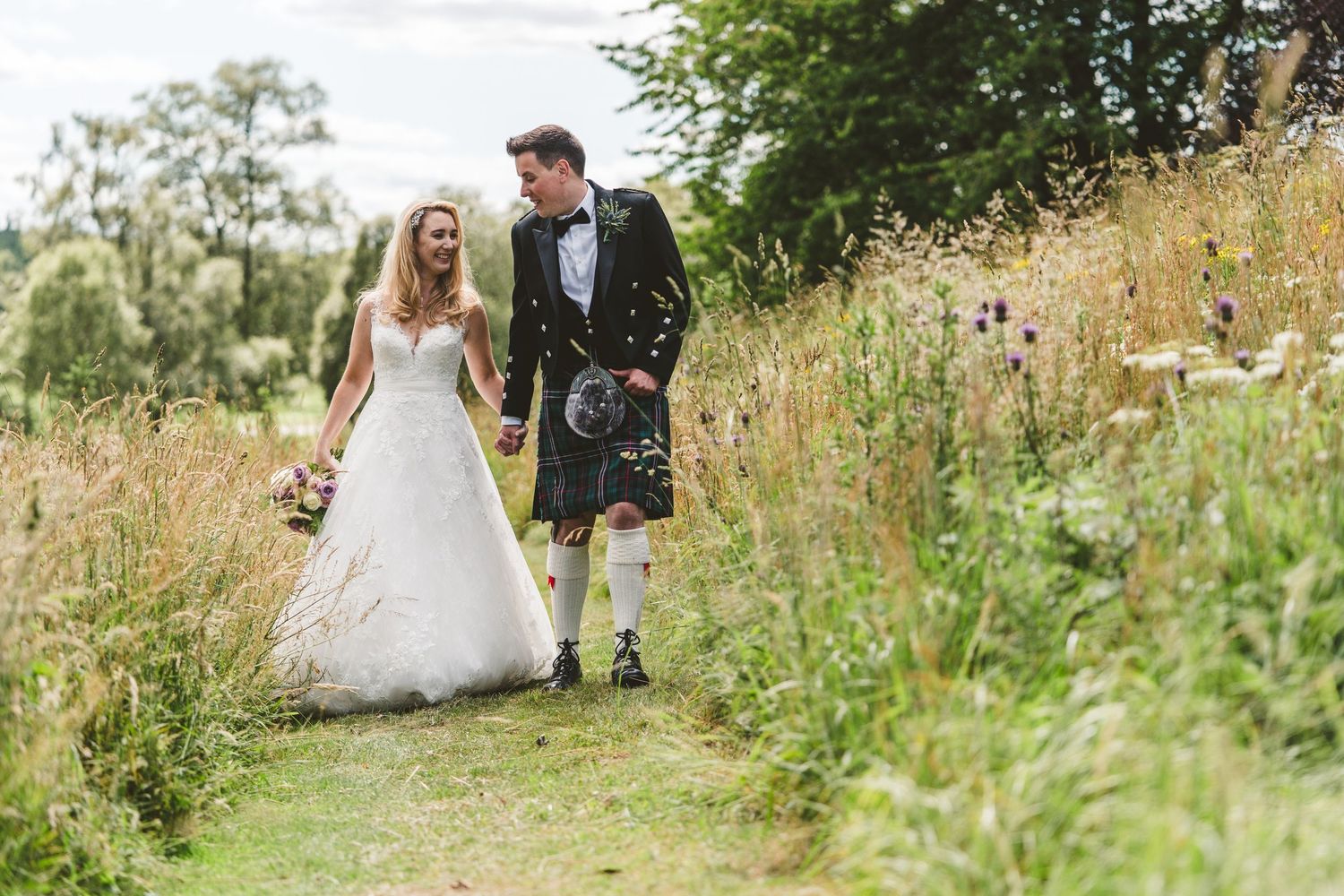 Wedding couple walks through tall summer grass in outdoor portrait session.